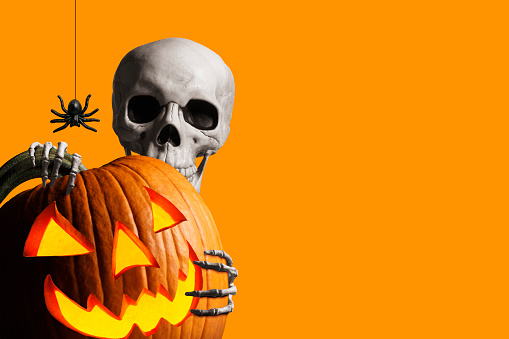 Skeleton peeks out from behind a jack olantern in front of orange background stock photo