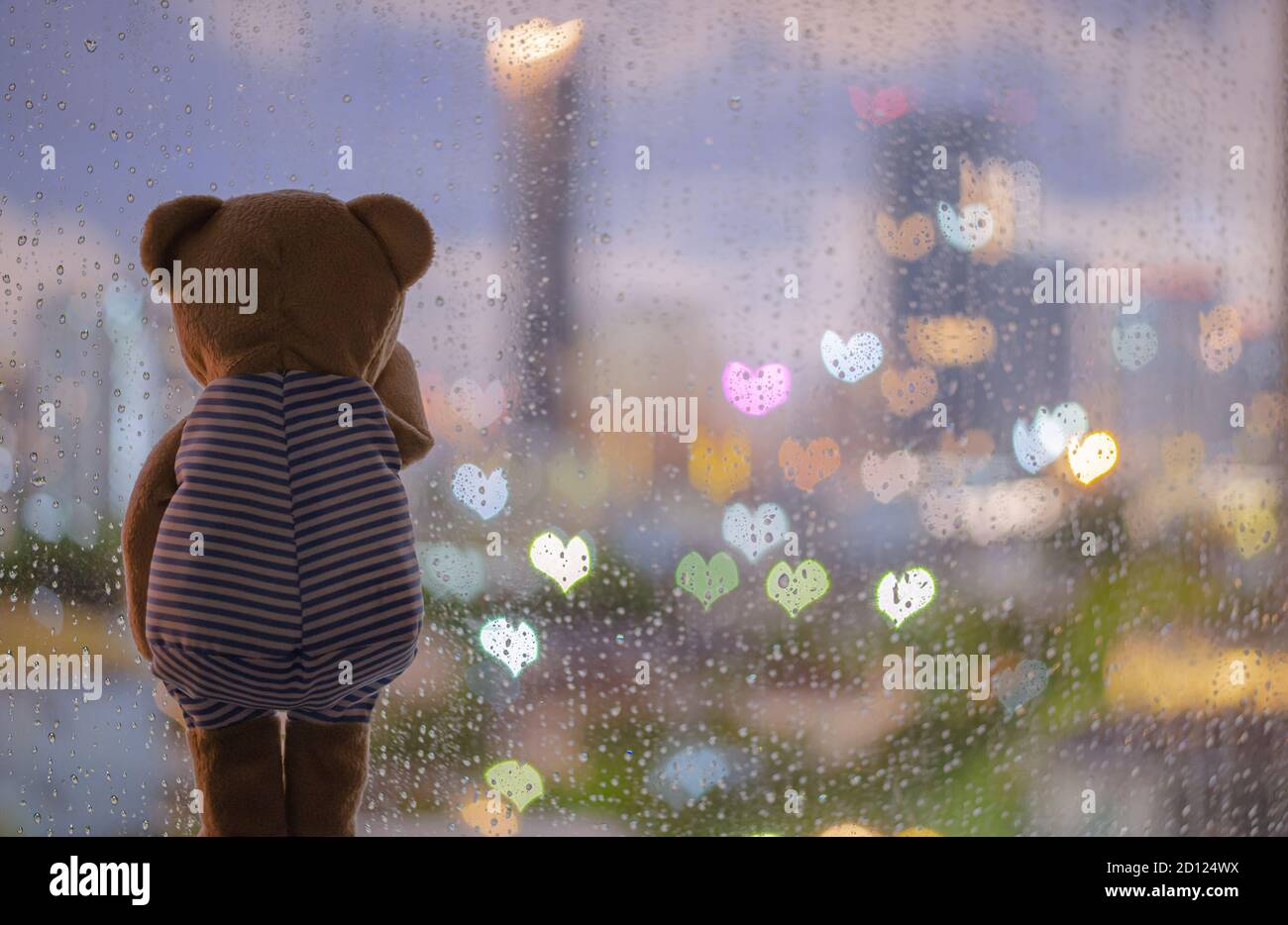 Teddy bear crying alone at window when raining with colorful love shape bokeh lights stock photo