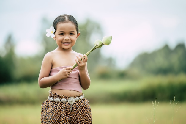 Free photo portrait of lovely little girls in thai traditional dress and put white flower on her ear standing and hold two lotus in hand on rice field she toothy smile
